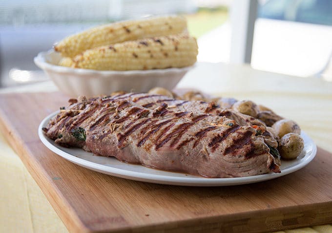 grilled stuffed pork tenderloin on a white plate on a cutting board with a bowl of grilled corn on the cob in the background