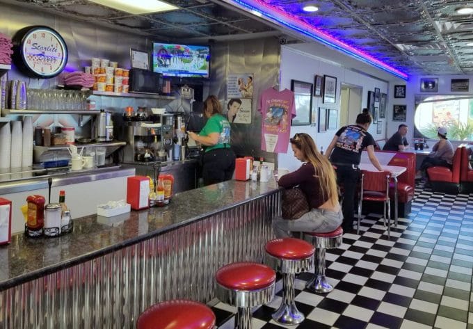 starlite diner in Daytona beach with black and white checkered floor, red top stools and a metal counter top with a woman sitting at the counter, booths in the background with people working