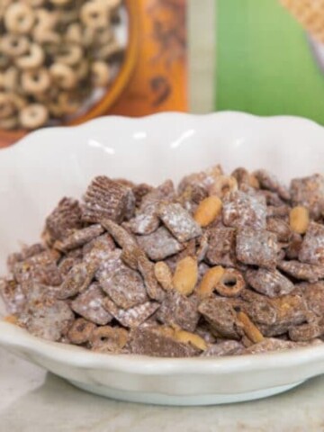 a close up shot of Chocolate Peanut Butter Cheerios and Chex Muddy Buddies in a white bowl with cereal boxes in the background on a kitchen counter top