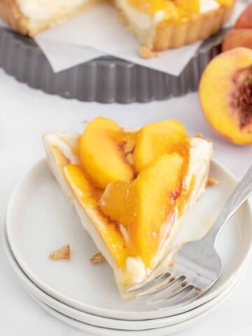 slice of peach tart on white plate with a fork with whole pie behind it