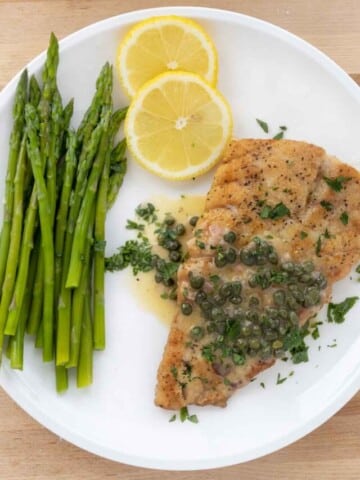 pan seared rockfish with a lemon caper sauce on a white plate with asparagus and lemon circles