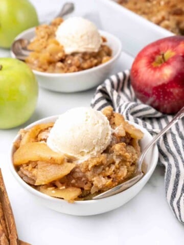 white bowl of apple crisp with ice cream next to whole red and green apples