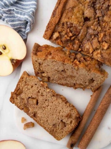 apple bread sliced with apples and cinnamon sticks on a white cutting board.