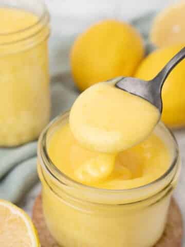 spoon in a jar of lemon curd, with lemons and another jar in the background.