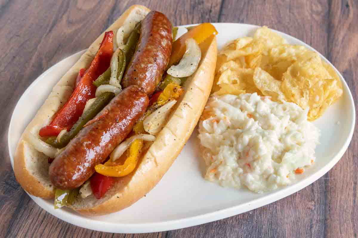 Smoked sausage, peppers and onions sandwich on a white platter with potato chips and coleslaw.