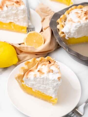 Slice of lemon meringue pie on a white plate with more pie in the background