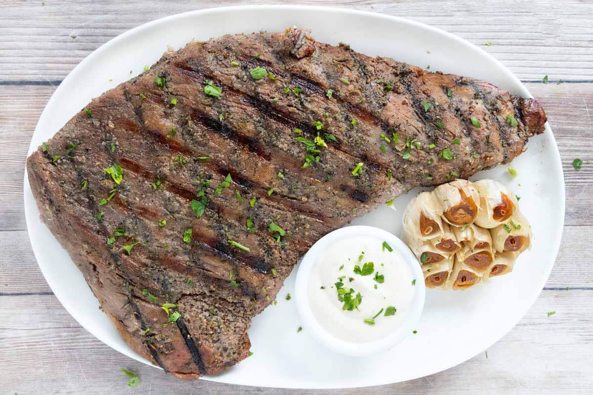 Smoked tri-tip steak on a white platter with smoked garlic and white sauce.