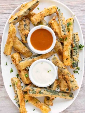 Fried zucchini sticks on a white platter with dipping sauces.