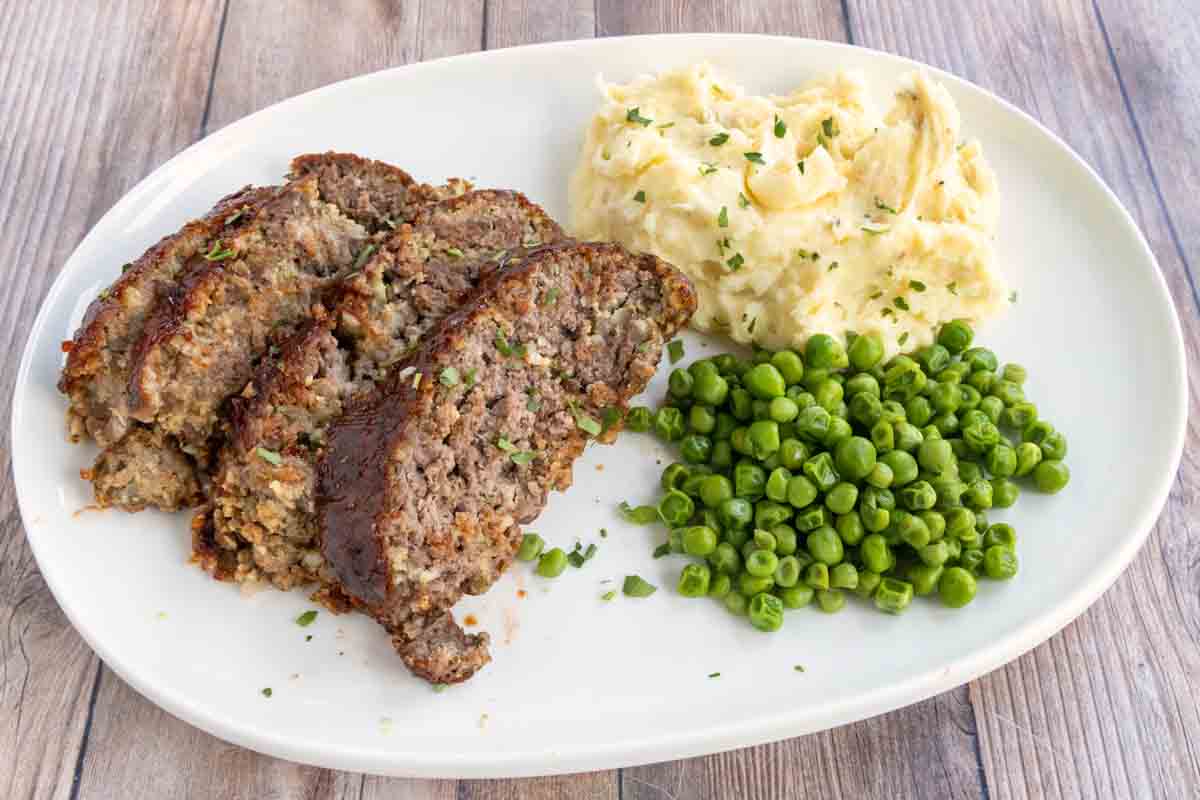 Sliced smoked meatloaf on a white plate with mashed potatoes and green peas.