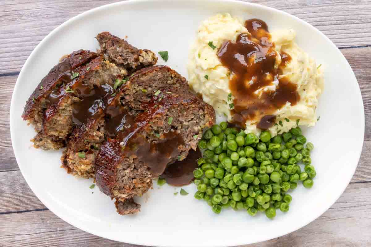 Smoked meatloaf with brown gravy, mashed potatoes and peas on a white plate.