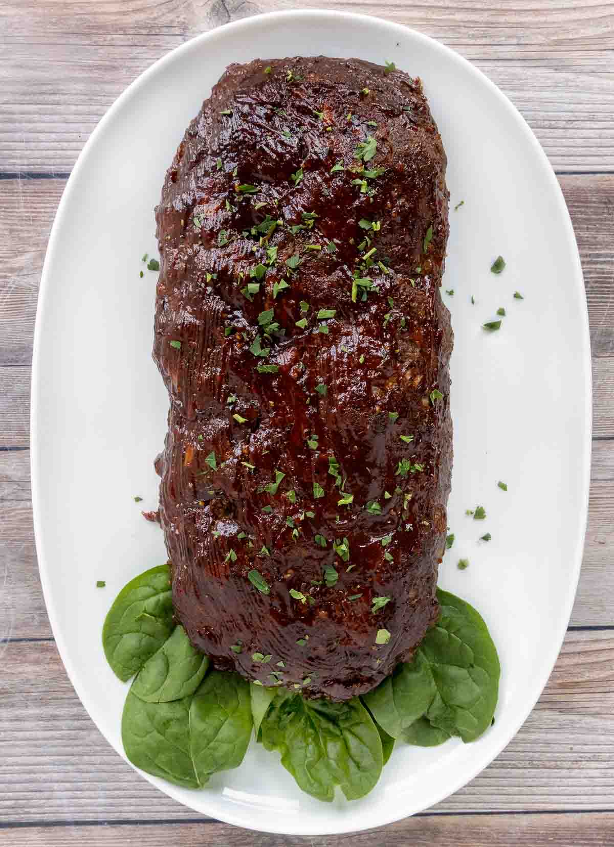 Smoked meatloaf on a white platter with spinach leaves.