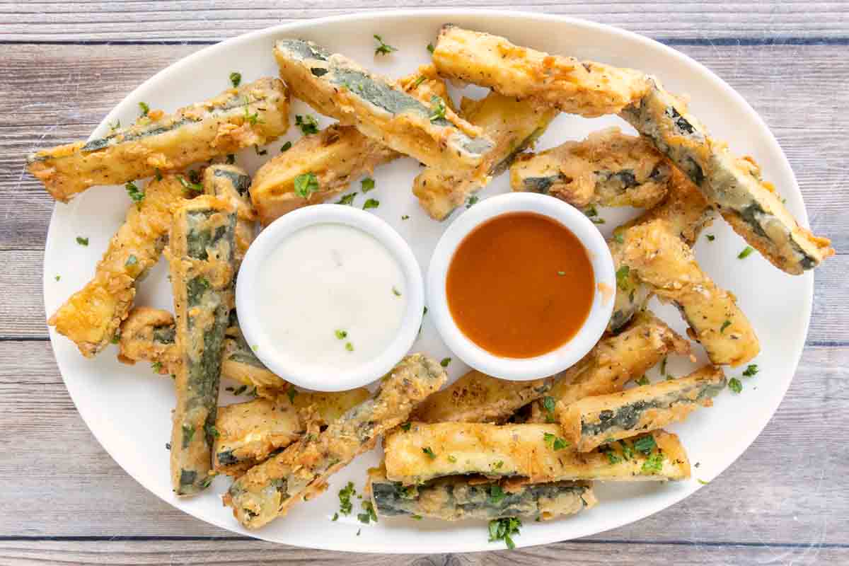 Fried zucchini sticks on a white platter with dipping sauces.