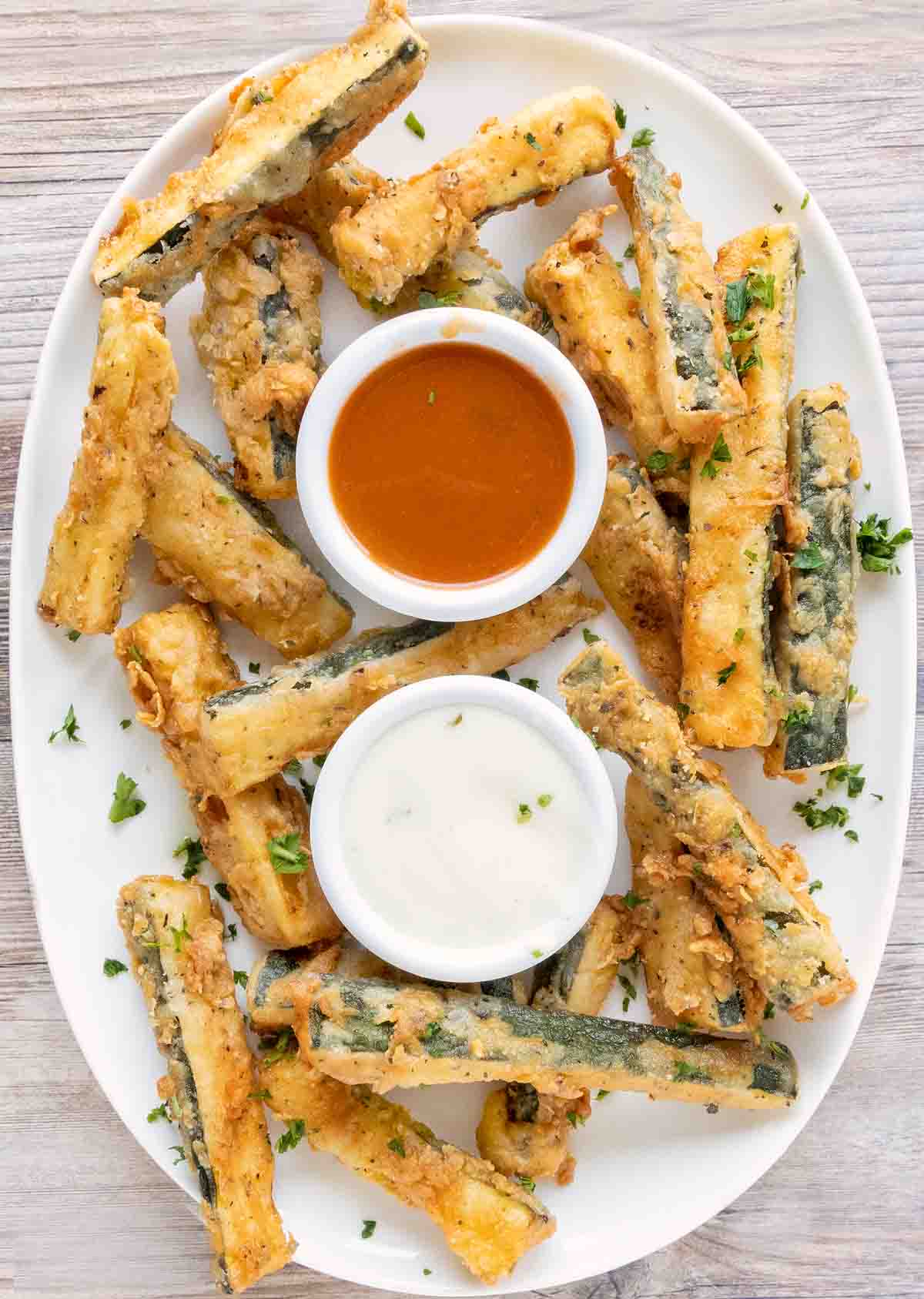 Fried zucchini sticks on a white platter with dipping sauces.