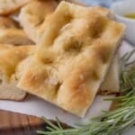 Slices of focaccia bread with sprig of rosemary on a brown cutting board.