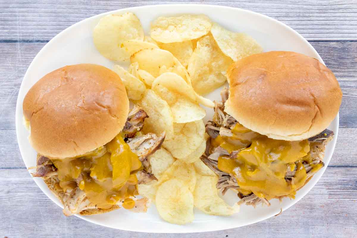 Two pulled pork sancdiches with carolina gold bbq sauce on a white plate with potato chips.