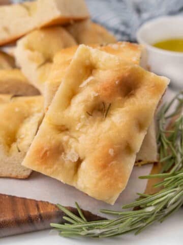 Slices of focaccia bread with sprig of rosemary on a brown cutting board.