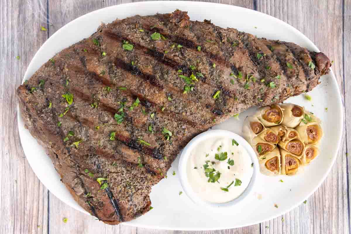 Tri-tip steak with smoked garlic and horseradish sauce on a white platter.