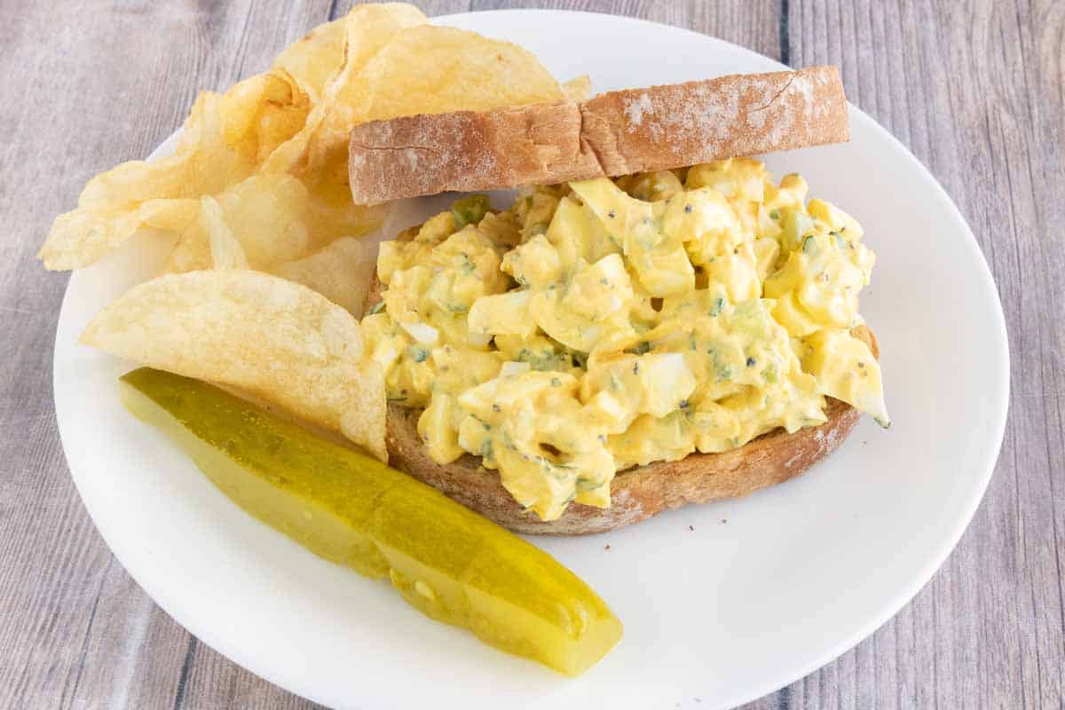 Egg salad sandwich on a white plate with potato chips and pickle spear.