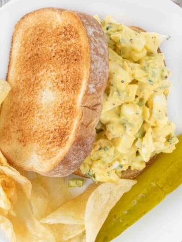 Egg salad sandwich on a white plate with potato chips and pickle spear.