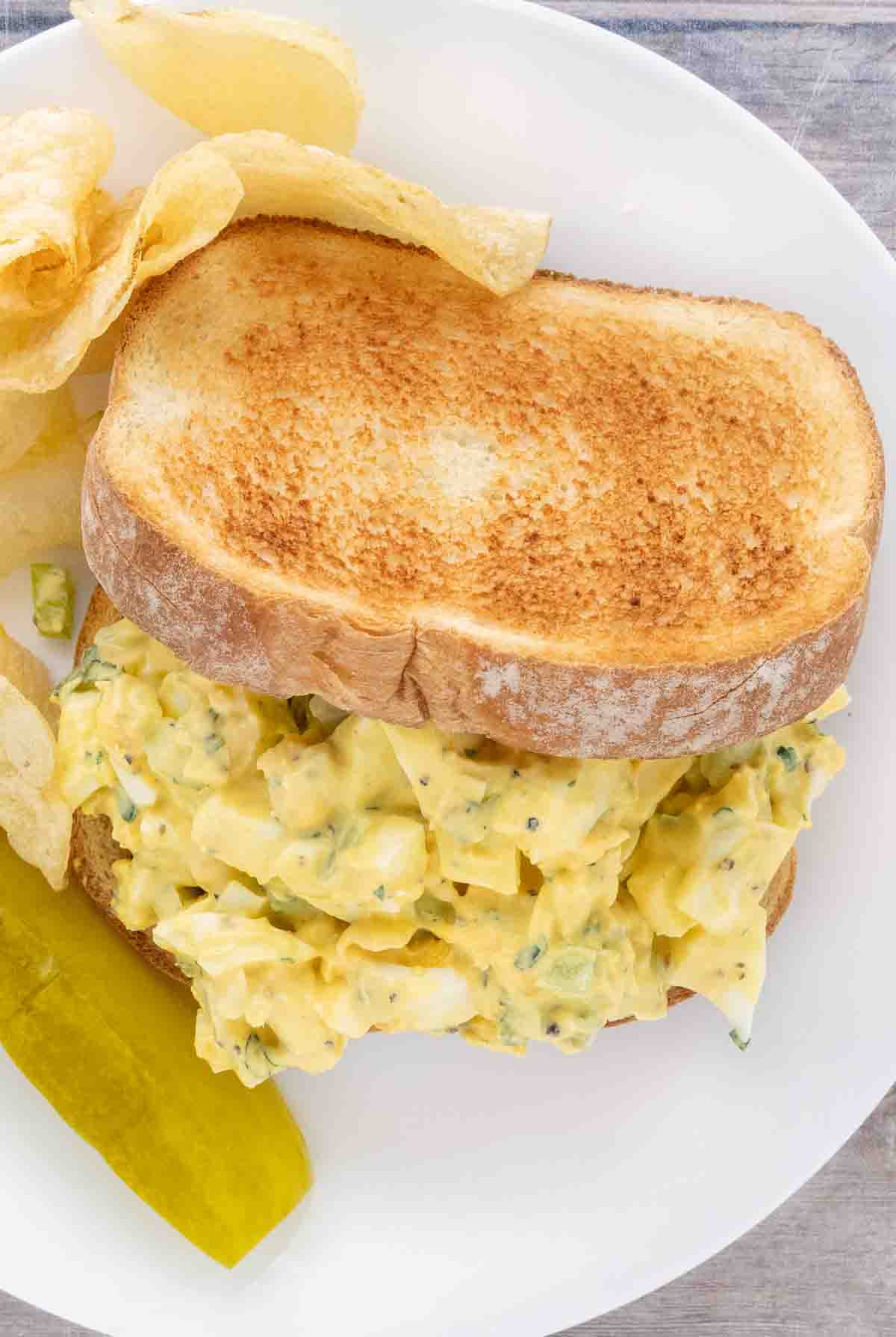 Egg salad sandwich on a white plate with potato chips and pickle spear.