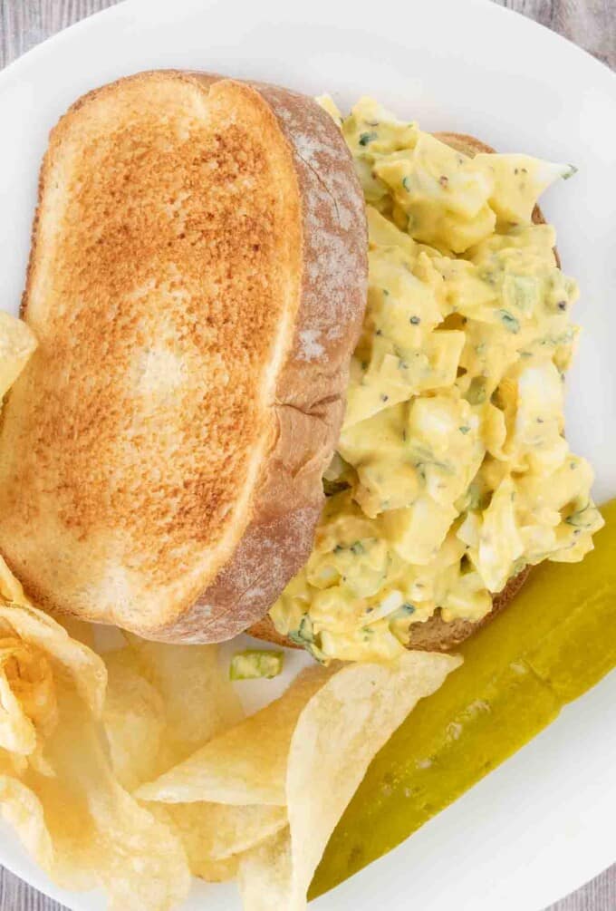 Egg salad sandwich on a white plate with potato chips and pickle spear.