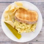 Egg salad sandwich on a white plate with potato chips and pickle spear.