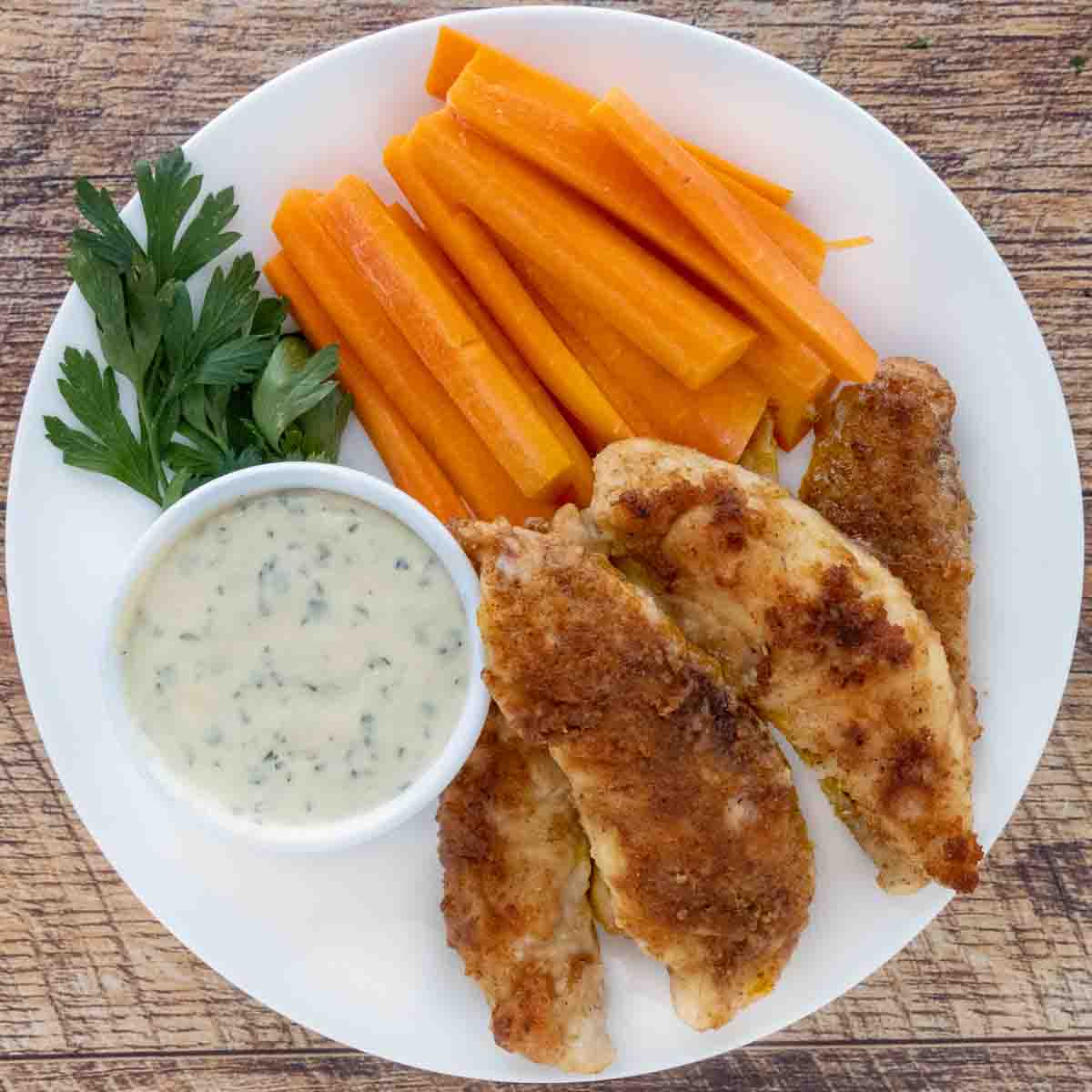 Chicken tenders and carrot sticks on a white plate with garlic parmesan sauce.