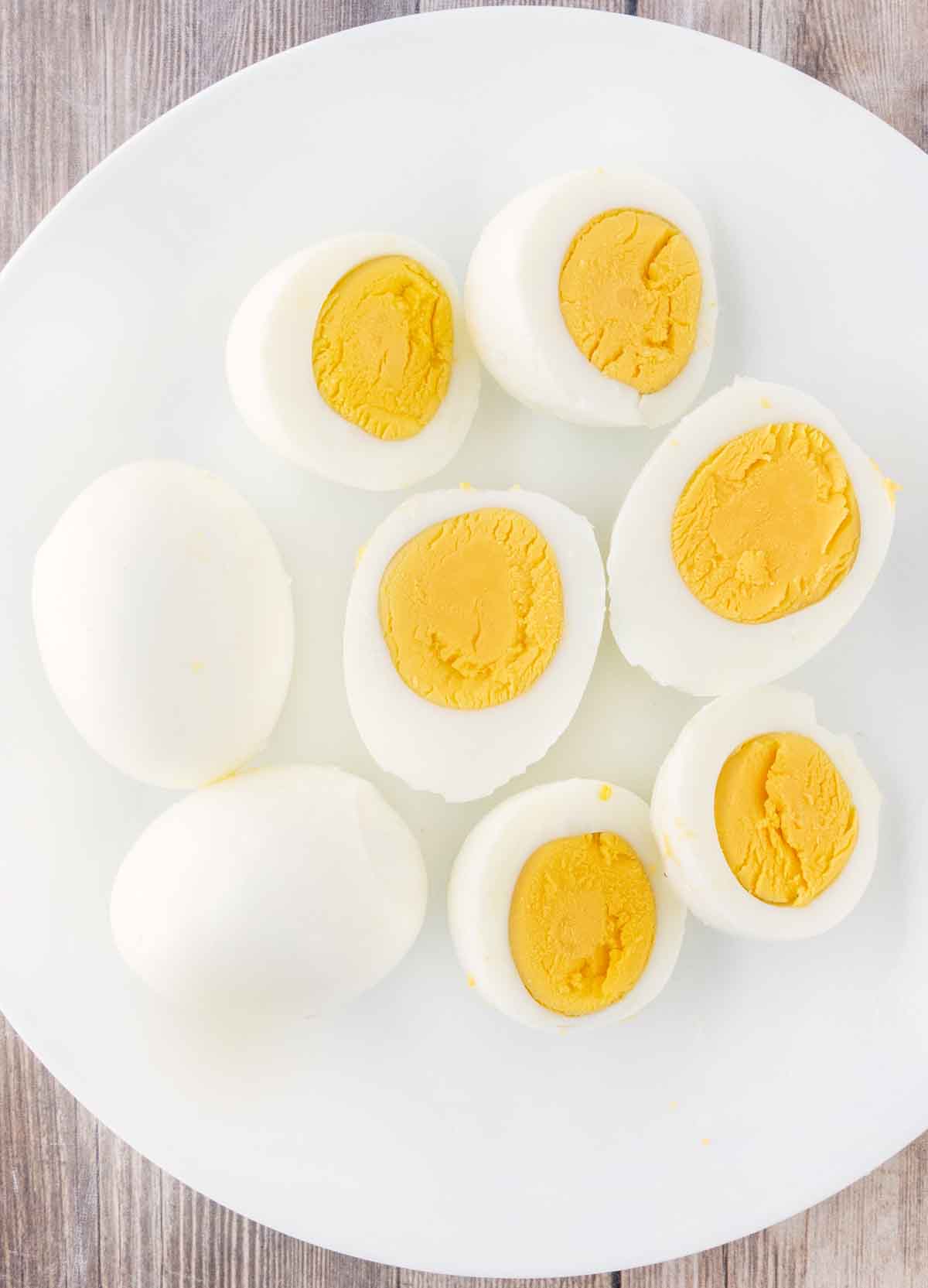 Two hard-boiled eggs next to eggs sliced in half on a white plate.