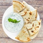 Raita sauce in a glass bowl on a white platter with pita bread.