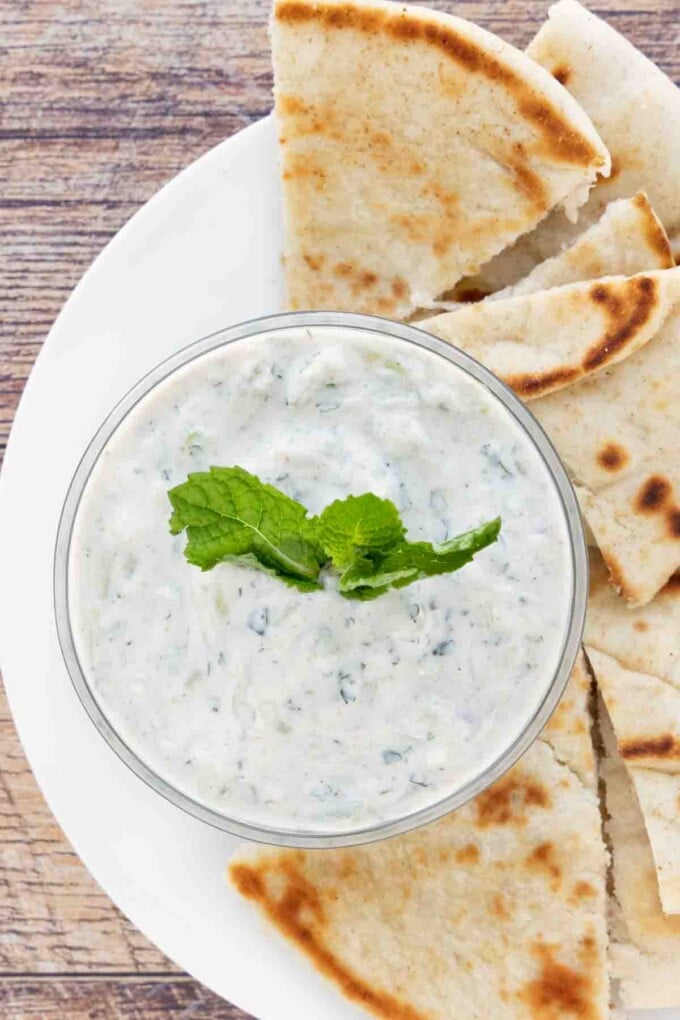 Raita sauce in a glass bowl on a white platter with pita bread.