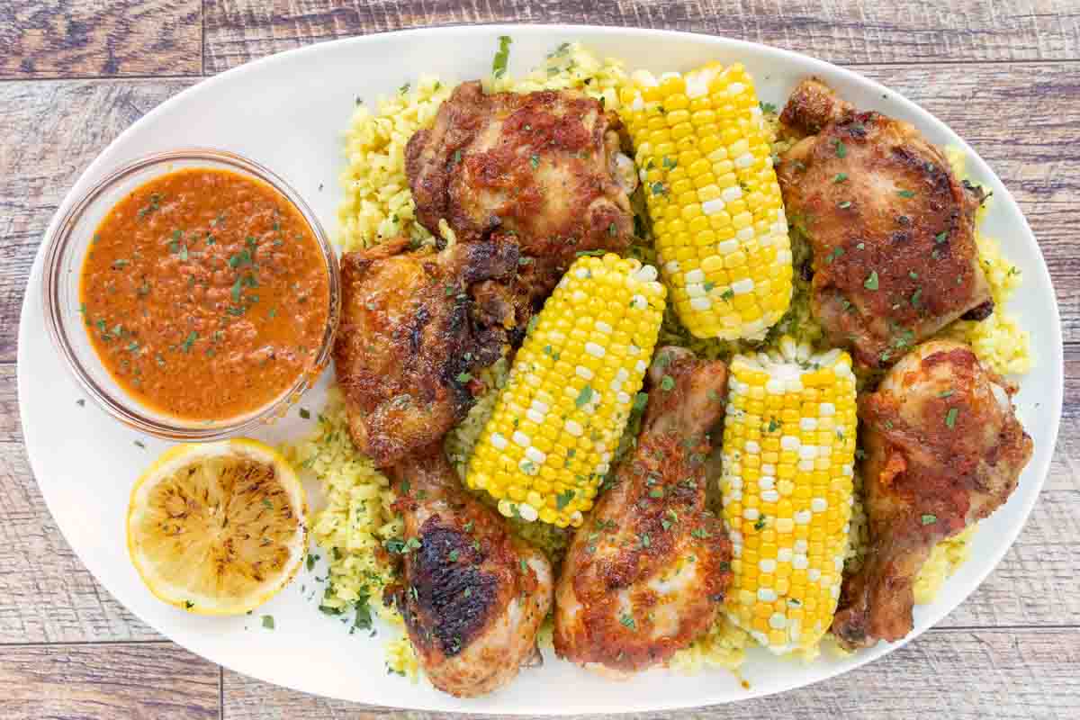 Per-peri chicken on a white platter with corn on the cob and peri peri sauce.