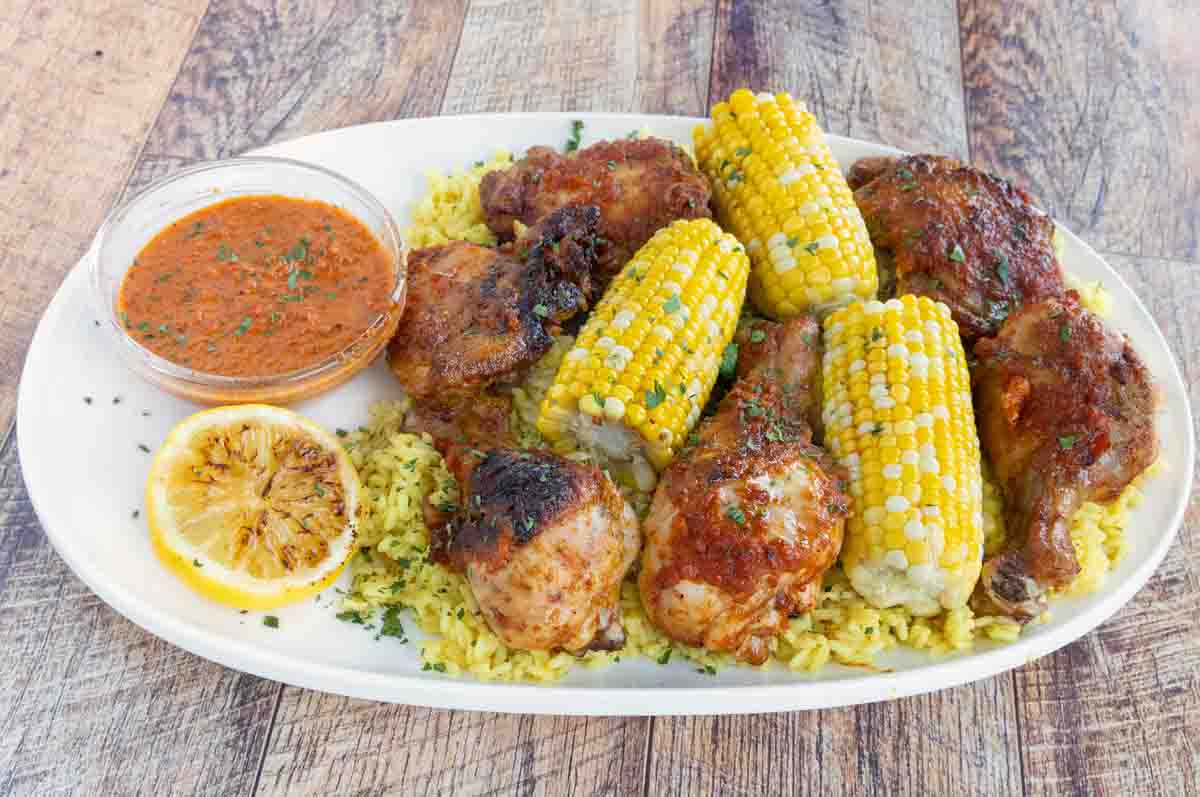 Per-peri chicken on a white platter with corn on the cob and peri peri sauce.