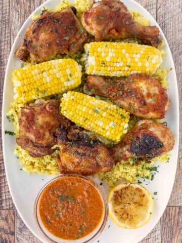 Per-peri chicken on a white platter with corn on the cob and peri peri sauce.