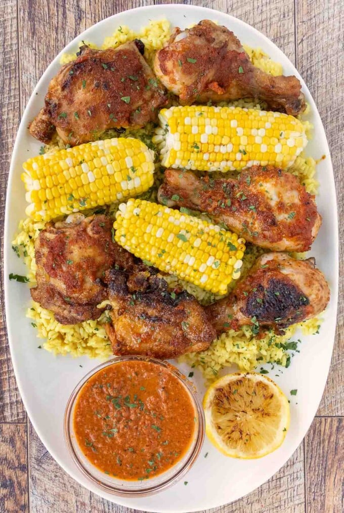 Per-peri chicken on a white platter with corn on the cob and peri peri sauce.