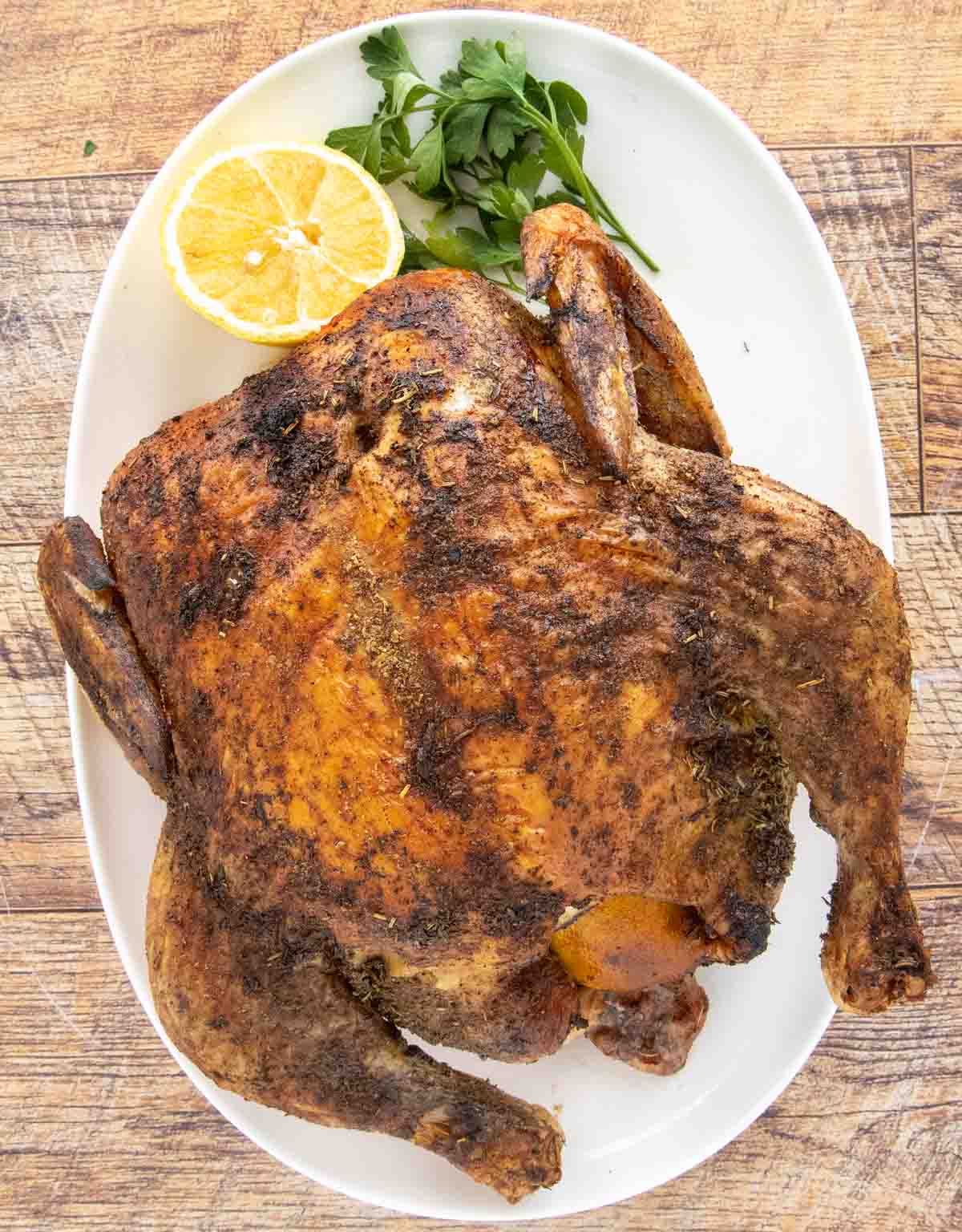 Whole herb roasted chicken on a white platter with a lemon half and parsley.