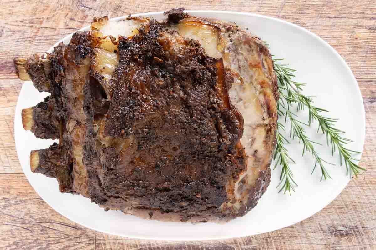 Whole cooked prime rib on a white platter with a sprig of rosemary.