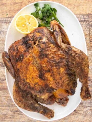 Whole herb roasted chicken on a white platter with a lemon half and parsley.