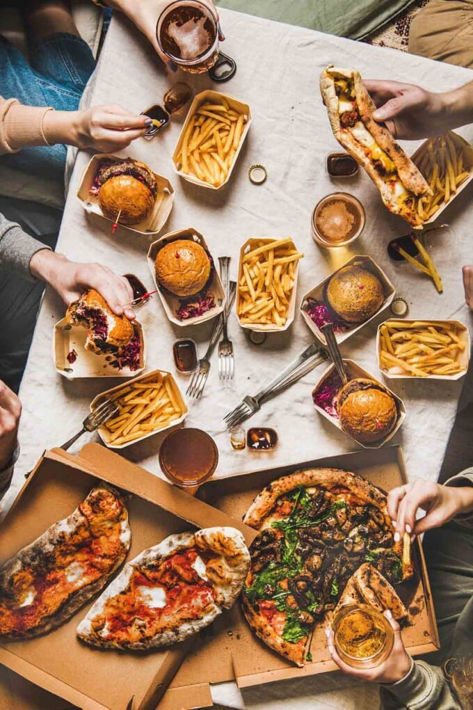 Table with pizza, burgers and fries with hands reaching for them.