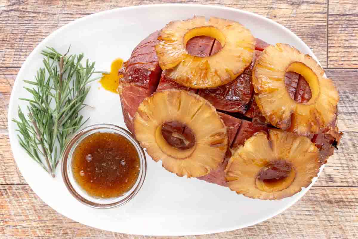 Whole smoked ham topped with pineapple with slices rosemary and glaze on a white platter.