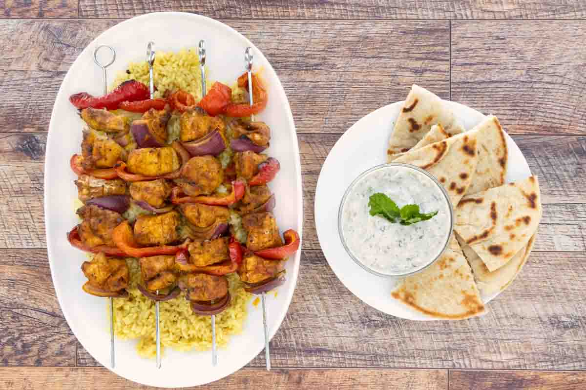 Skewered chicken kabobs on a bed of yellow rice on a white platter next to a platter of sauce and pita.