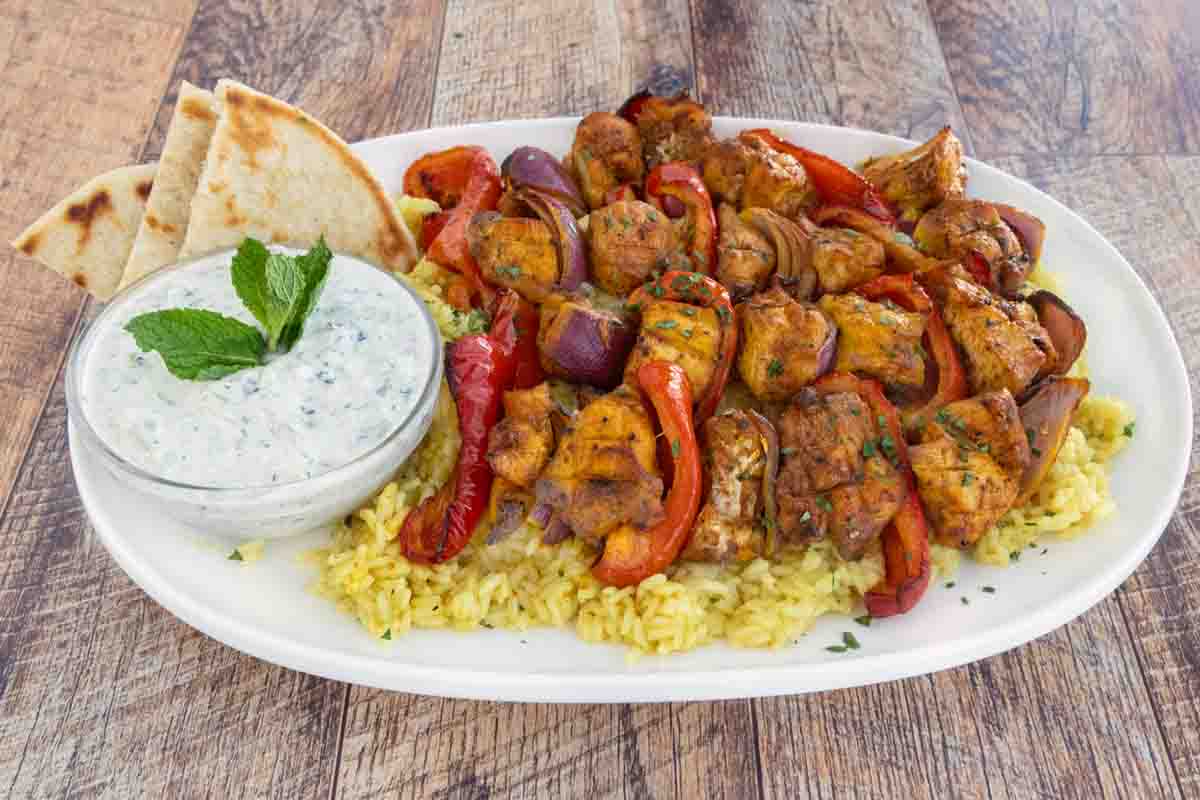 Chicken kabobs on a bed of yellow rice with sauce and pita on a white platter.