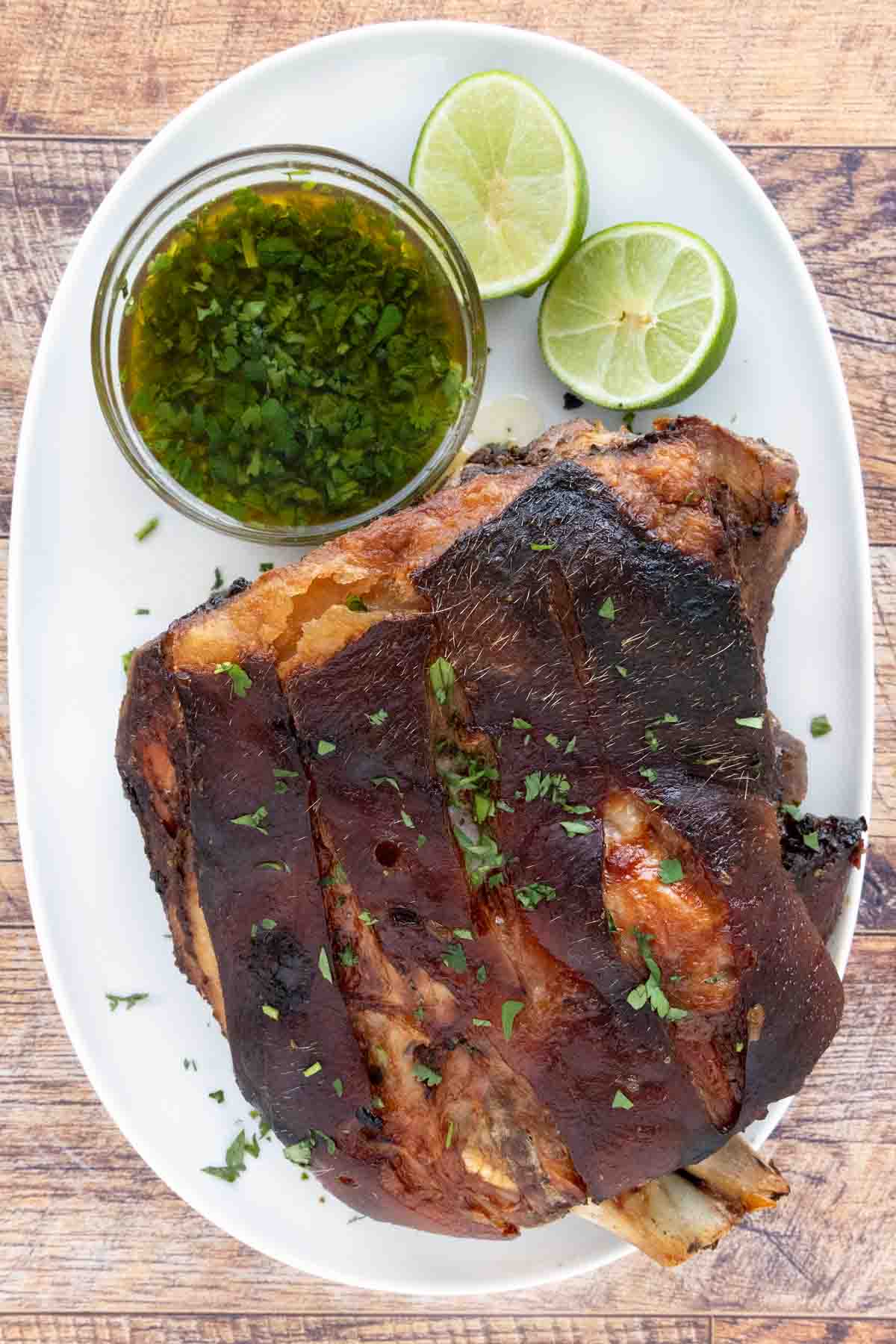 Pernil (Puerto Rican roast pork) on a white platter with green sauce and limes.