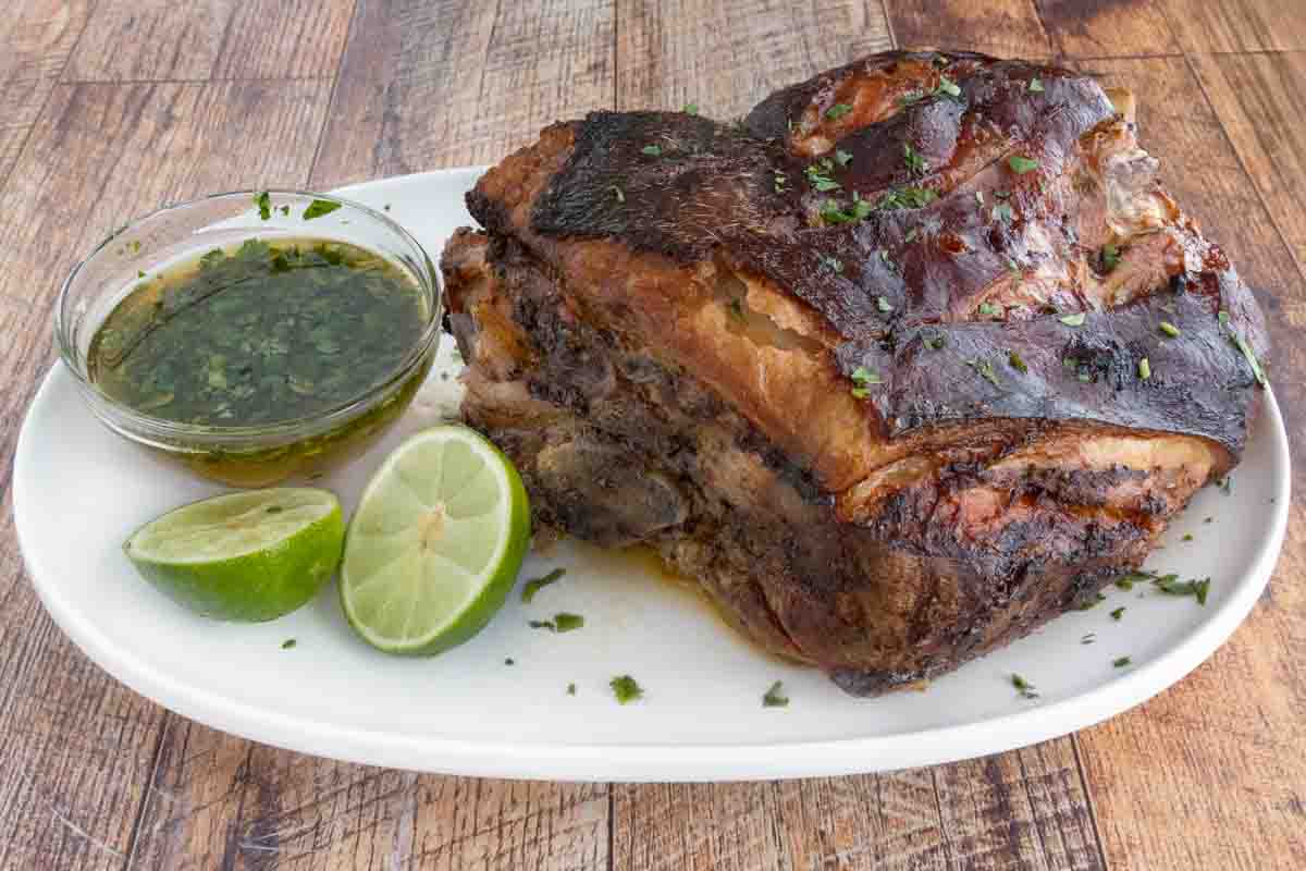 Pernil (Puerto Rican roast pork) on a white platter with green sauce and limes.