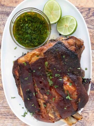 Pernil (Puerto Rican roast pork) on a white platter with green sauce and limes.