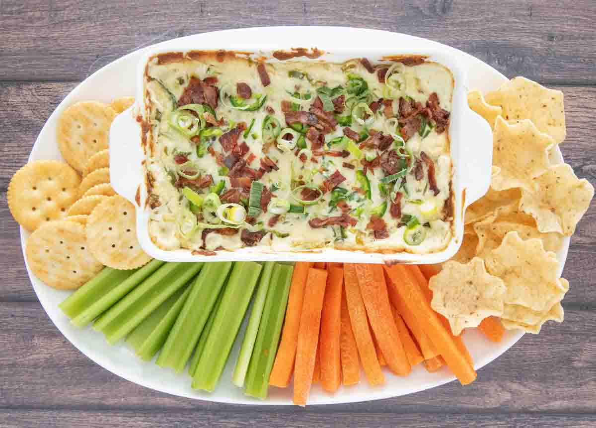 Baked jalapeno popper dip in a white casserole with carrots, celery and chips on a white platter.