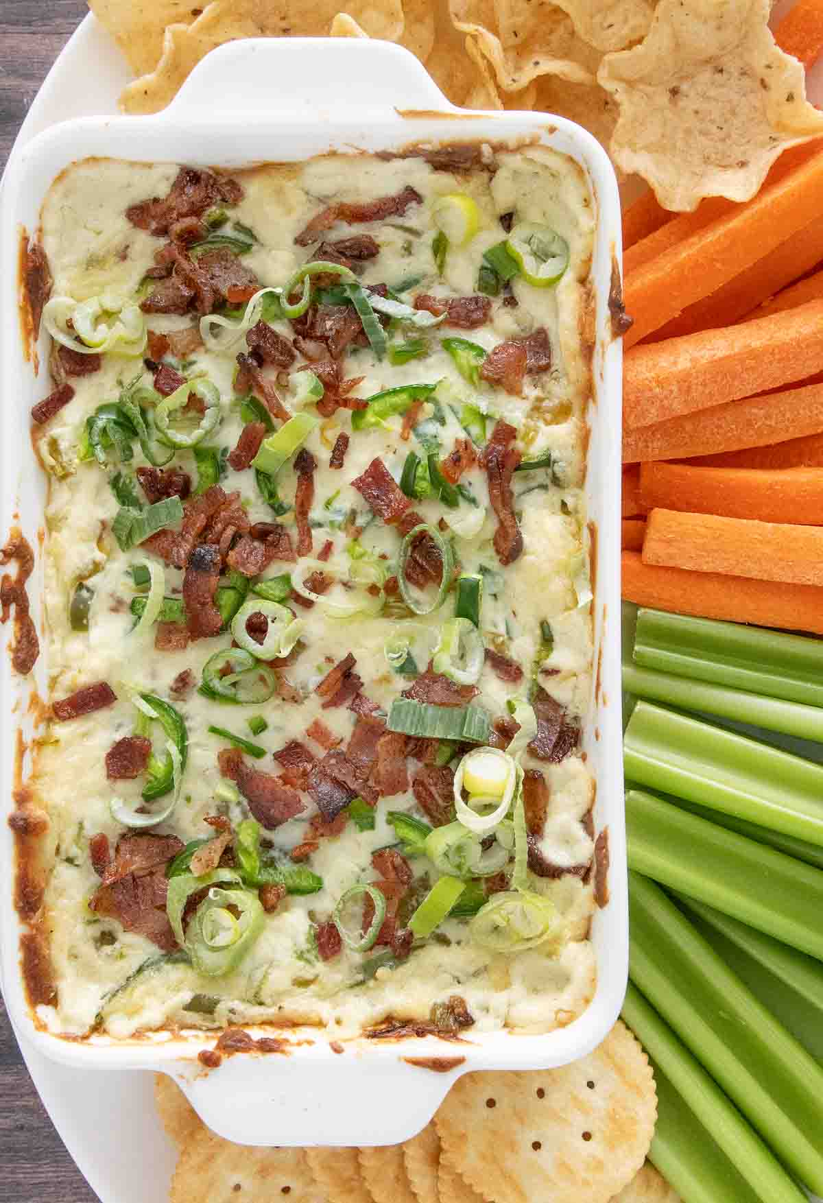 Baked jalapeno popper dip in a white casserole with carrots, celery and chips on a white platter.