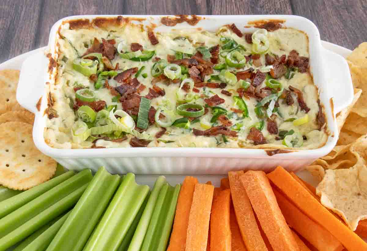 Baked jalapeno popper dip in a white casserole with carrots, celery and chips on a white platter.