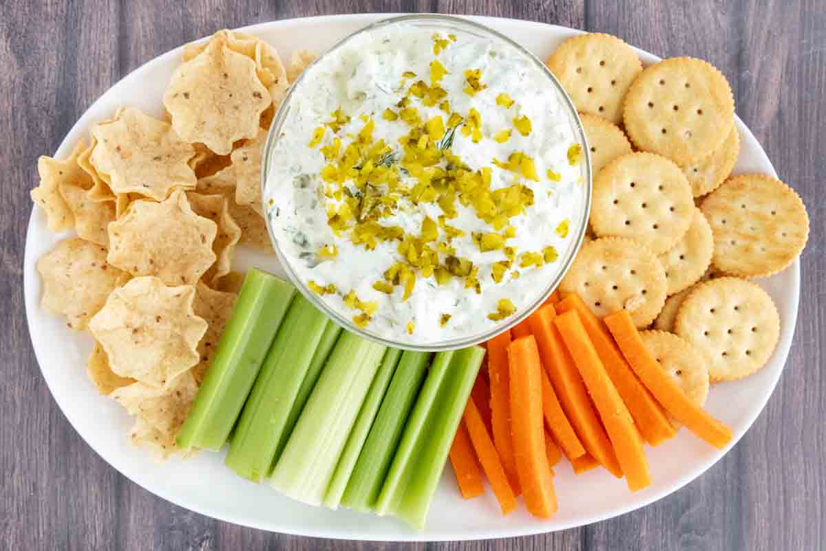 Dill pickle dip a white bowl on a platter with crackers, tortilla chips, celery sticks and carrot sticks.