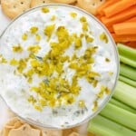 Pickle dip a white bowl on a platter with crackers, tortilla chips, celery sticks and carrot sticks.