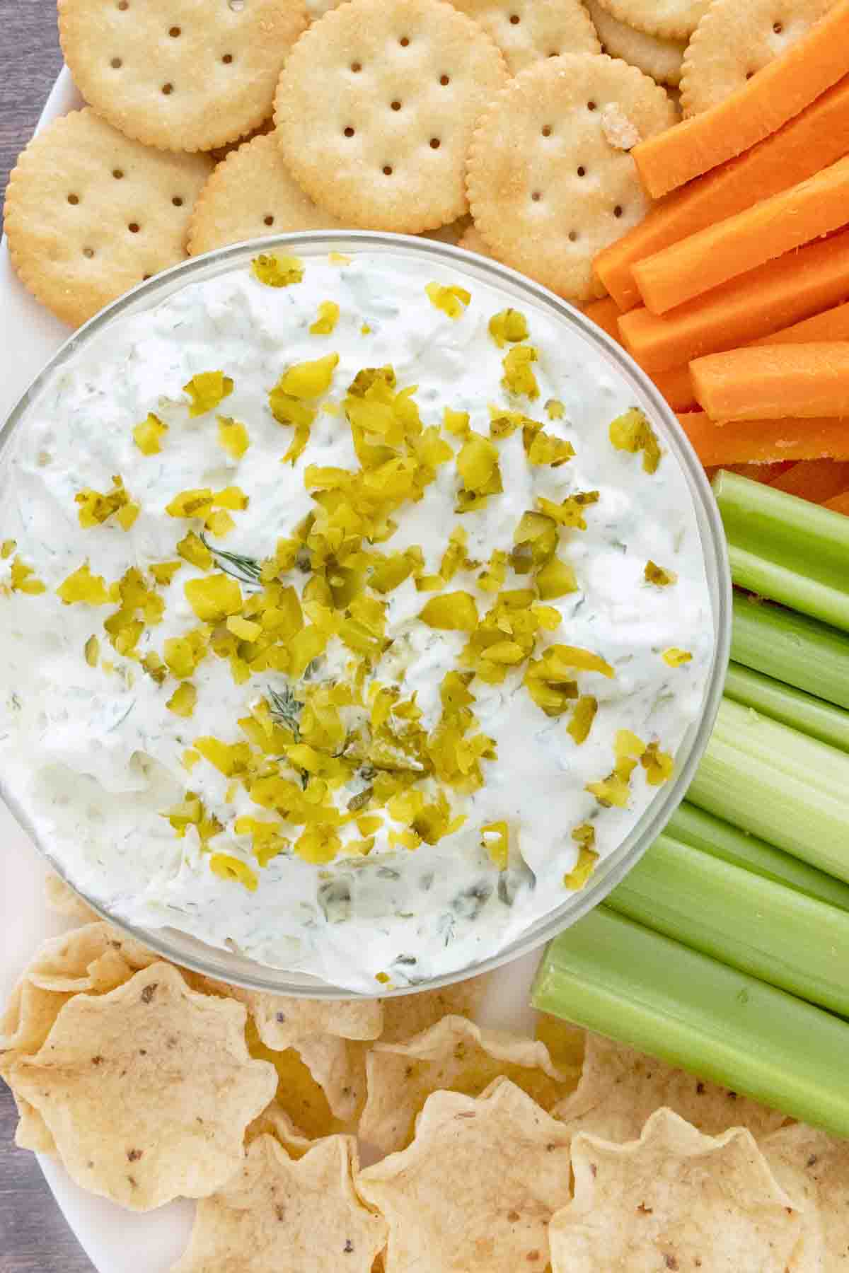 Dill Pickle dip a white bowl on a platter with crackers, tortilla chips, celery sticks and carrot sticks.